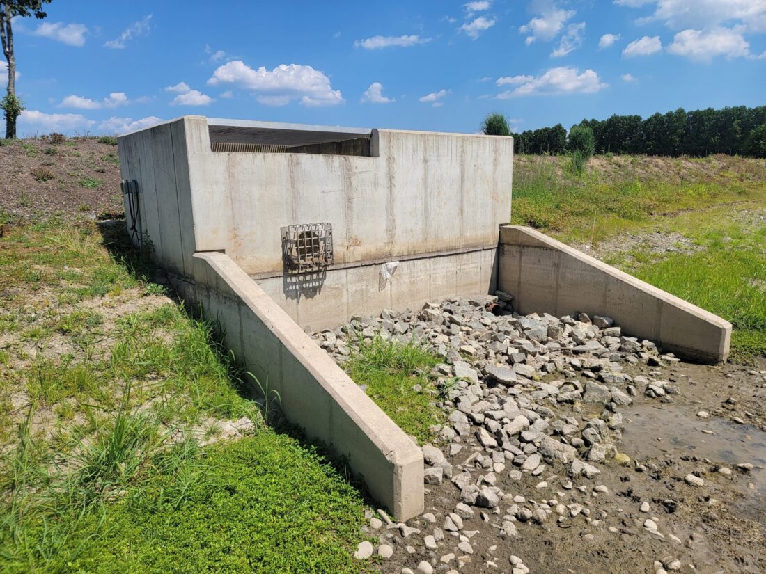 Concrete stormwater outlet control structure with surrounding rocks and vegetation, designed to manage stormwater discharge and prevent erosion.